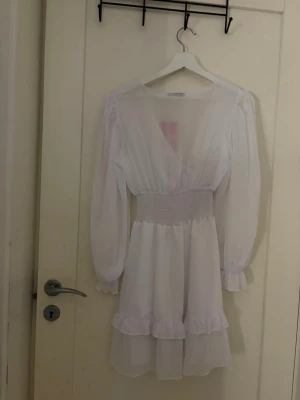 Vit kortklänning med volang och knyt - Söt vit kortklänning med omlottdesign och knyt framtill. Klänningen har långa ärmar med resår vid handlederna, markerad midja med smock och volangdetaljer längst ner. Perfekt för sommarens alla tillfällen.helt ny aldrig använden 