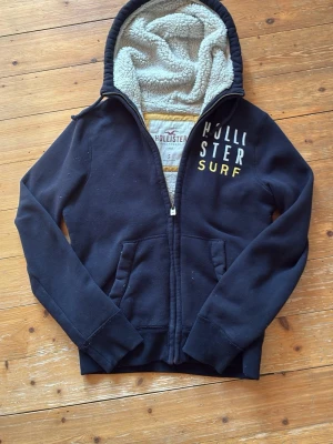 Svart Hollister hoodie med teddyfoder - Mysig svart hoodie från Hollister med dragkedja och gul/vit text på bröstet. Insidan och huvan är fodrade med vitt teddyfoder för extra värme. Två fickor framtill och ribbade muddar vid ärmslut och nederkant. Perfekt för kalla dagar.