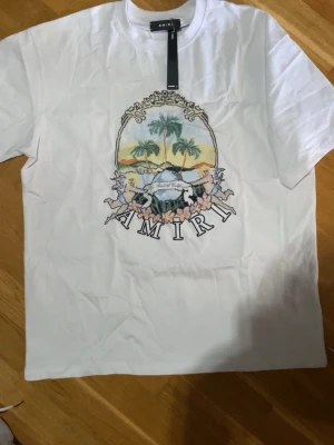Vit t-shirt med tryck från Amiri - Säljer en vit t-shirt från Amiri med stort färgglatt tryck framtill som visar palmer, solnedgång och texten 'AMIRI'. T-shirten är i 100% bomull och har klassisk passform med korta ärmar. Perfekt för dig som gillar streetwear och snygga prints.