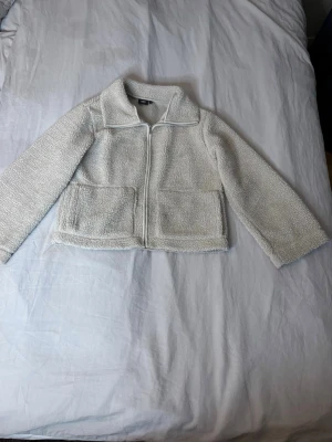 Ljus beige teddyjacka med dragkedja - Mysig vit teddyjacka i fluffigt material med dragkedja framtill och två stora fickor. Jackan har en klassisk krage och raka ärmar. Perfekt för att hålla värmen under kyliga dagar och ger en trendig look.
