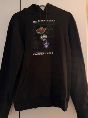 Svart NHL Global Series hoodie - Säljer en svart hoodie med tryck från NHL Global Series Sweden 2023. Framsidan har loggor för Minnesota Wild och Toronto Maple Leafs samt text och grafik. Perfekt för dig som gillar hockey och sportig stil.