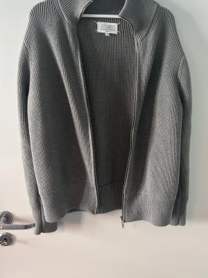 Grå maison margiela zip  - Också Köpt på vinted. Snygg grå ribbstickad tröja med dragkedja framtill och hög krage. Tröjan har långa ärmar och breda muddar vid ärmslut och nederkant. Perfekt för lager på lager och enkel att styla till olika outfits.