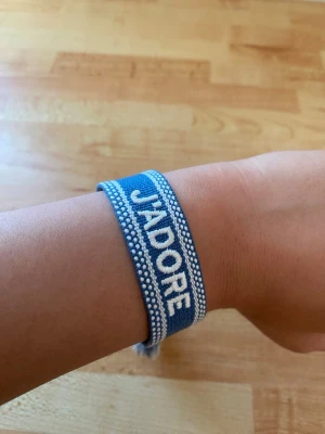 Blått tygarmband med text - Snyggt blått tygarmband med vita detaljer och texten 'J'ADORE' broderat på framsidan. Armbandet har justerbara knytband med tofsar i ändarna och ett mönster av små vita prickar längs kanterna. Köpt från Frankrike! Inte använd! vV sälja eftersom använder aldrig.