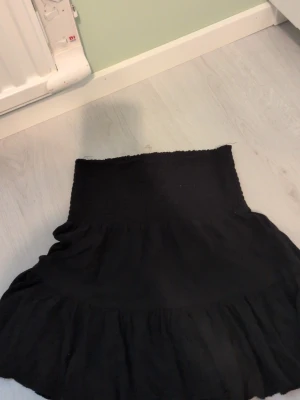 Svart volangkjol XS - Supersöt svart kjol i storlek XS med bred smock i midjan och volang nedtill. Kjolen är kort och har en enkel, stilren look som passar perfekt till många olika outfits.