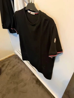 Svart t-shirt med Moncler-detalj - Snygg svart t-shirt med rund hals och korta ärmar. Ärmsluten har rödvita ränder och på vänster ärm sitter Moncler-loggan. Perfekt till jeans eller shorts för en clean och stilren look. PRIS kan diskuteras !