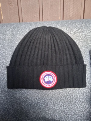 Svart ribbstickad mössa Canada Goose - Säljer en svart ribbstickad mössa från Canada Goose med uppvikt kant och det klassiska runda röda och vita märket framtill. Perfekt för kalla dagar och riktigt snygg till alla vinteroutfits.