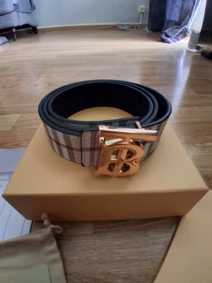 Rutig Burberry bälte med guldspänne - Snyggt bälte från Burberry med klassiskt rutigt mönster i beige, svart och rött. Bältet har ett stort guldfärgat spänne med Burberrys ikoniska TB-logga. Perfekt accessoar för att lyfta vilken outfit som helst.