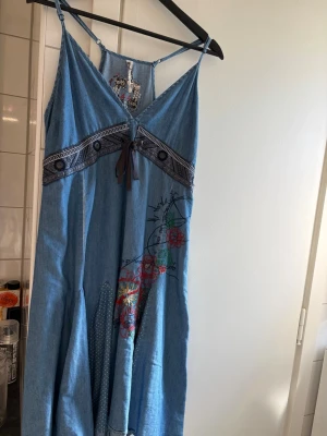 Blå långklänning från Desigual - Unik blå långklänning från Desigual i jeansmaterial med broderade blommor och dekorativ snörning framtill. Klänningen har smala axelband och v-ringning, samt detaljer i form av band och broderier som ger en bohemisk känsla.