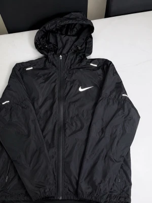 Svart Nike vindjacka med huva - Snygg svart vindjacka från Nike med vit logga på bröstet och reflekterande detaljer. Jackan har dragkedja framtill, två sidofickor med dragkedja och justerbar huva. Tillverkad i lätt och tåligt material, perfekt för blåsiga dagar.