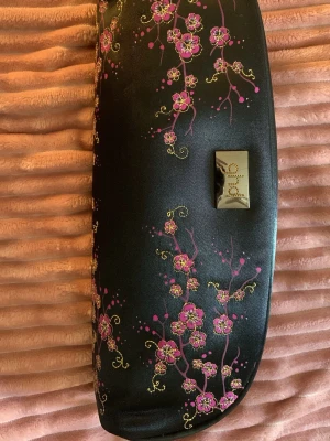 Svart blommigt fodral från ghd - Snyggt svart fodral från ghd med blommigt mönster i rosa och guld. Fodralet har en elegant form och ett metallmärke framtill. Perfekt för att förvara stylingverktyg eller skönhetsprodukter. Materialet är glansigt och känns lyxigt. Kan tänka mig att gå ner lite i pris vid snabb & smidig affär!