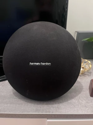 Harman Kardon rund bärbar högtalare - Snygg och stilren bärbar högtalare från Harman Kardon i svart tygmaterial. Den har en rund, modern design som passar perfekt i hemmet eller på resan. Högtalaren levererar kraftfullt ljud och har en tydlig logga på framsidan.