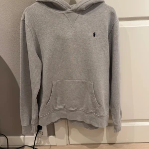 Grå hoodie Polo Ralph Lauren - Klassisk grå hoodie från Polo Ralph Lauren med broderad logga på bröstet. Tröjan har huva, magficka och långa ärmar. Tillverkad i mjuk bomullsblandning som är perfekt för kyligare dagar. Den har några fläckar på huvan, därför säljes billigare.