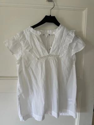 Vit blus med volangdetaljer - Söt vit blus med korta ärmar och volangdetaljer vid axlar och halsringning. Blusen har kvar prislapp o är därför aldrig använd.