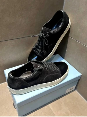 Svarta sneakers med mockadetaljer - Snygga svarta sneakers med ovandel i mocka och tåparti i blankt material. Skorna har klassisk snörning och vit sula som ger en clean kontrast. Perfekta för dig som gillar stilrena och tidlösa sneakers med en twist.