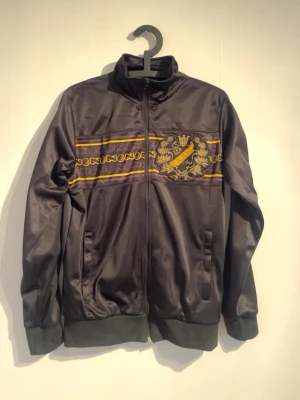 Svart AIK ziptröja med emblem - Snygg svart ziptröja med AIK:s klubbemblem broderat på bröstet och dekorativa gula och lila ränder över bröstet. Tröjan har hög krage, muddar vid ärmslut och nederkant samt två fickor framtill. Perfekt för dig som vill visa ditt AIK-stöd.