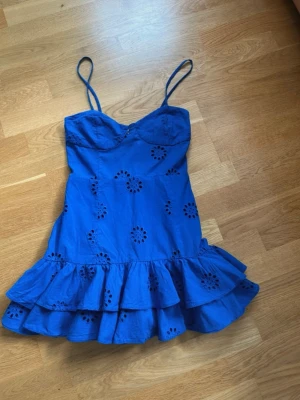  Trendig Blå kort klänning med volang - Supersöt blå klänning med smala axelband och volangkant nertill. Klänningen har broderade hålmönster i blommig stil och en figurnära passform upptill. Perfekt för varma dagar och sommarens alla äventyr.