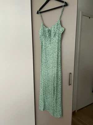 Grön blommig långklänning med slits - Snygg ljusgrön långklänning med små blommor, smala axelband och söt knytning framtill. Använd en gång, storlek M
