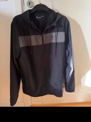 Svart vindjacka med huva Under Armour - Snygg svart vindjacka från Under Armour med grått bröstparti och halv dragkedja. Jackan har huva med dragsko och är tillverkad i lätt, vindtätt material. Perfekt för blåsiga dagar och sportiga outfits.