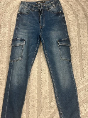 Blå cargobyxor i denim, storlek S - Snygga blå cargobyxor i denim med raka ben och stora fickor på sidorna. Byxorna har klassisk jeanslook med bälteshällor och knappstängning. Perfekta för en avslappnad streetstil och passar till många olika outfits.