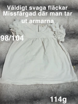 Vit topp med volangärmar - Supersöt vit topp med korta volangärmar och rund hals. Den har en lös och luftig passform som ger en chill vibe. Perfekt till jeans eller kjol för en enkel men söt look.🌸Svaga fläckar. Missfärgad där man tar ut armarna. Storlek 98/104