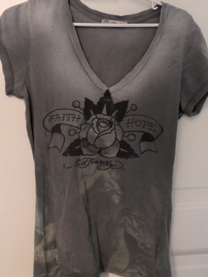 Grå Ed Hardy t-shirt med tryck - Snygg grå t-shirt från Ed Hardy med v-ringning och korta ärmar. Framsidan har ett stort tryck med ros och texten 'Faith Hope', medan baksidan pryds av en pegasus och Ed Hardy-logga. Perfekt för dig som gillar streetwear och unika prints. Den är vintage och köppt i Usa under 2000 talet. 