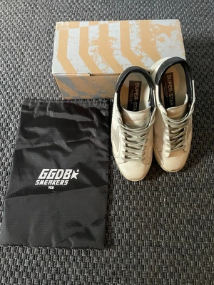 Golden Goose skor - Snygga Golden Goose GGDB Superstar sneakers i vitt läder med den ikoniska stjärnan på sidan och svarta detaljer vid hälen. Skorna har gråvita skosnören och den klassiska slitna stilen. Kommer med originalkartong och dustbag. Perfekta för en trendig och avslappnad stil.