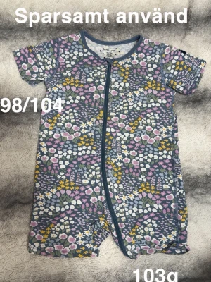 Blommig kortärmad pyjamas med dragkedja - Kortärmad pyjamas med färgglatt blommönster i lila, blått, vitt och gult. Pyjamasen har dragkedja framtill och korta ben, vilket gör den både praktisk och bekväm. Perfekt för mysiga kvällar och sommarens varma nätter.🌸 Sparsamt använd. 98/104