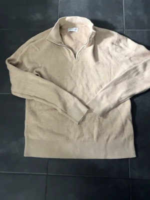 Beige halv zip tröja These Glory Days - Snygg beige stickad tröja från These Glory Days i storlek S. Tröjan har halvzip, hög krage och är ribbstickad med långa ärmar. Perfekt för lager på lager och enkel att matcha med olika outfits.