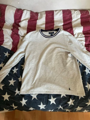 Grå långärmad tröja Polo Ralph Lauren - Mysig grå långärmad loungewear-tröja från Polo Ralph Lauren. Tröjan har rund halsringning med marinblå och vit rand, samt den klassiska lilla loggan broderad nere vid fållen. Perfekt för chill dagar hemma.
