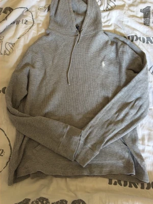 Grå hoodie från Polo Ralph Lauren - Säljer en grå hoodie från Polo Ralph Lauren i mjukt, strukturerat bomullstyg. Klassisk huva med dragsko, långärmad modell och liten vit logga broderad på bröstet. Perfekt för en avslappnad och stilren look.
