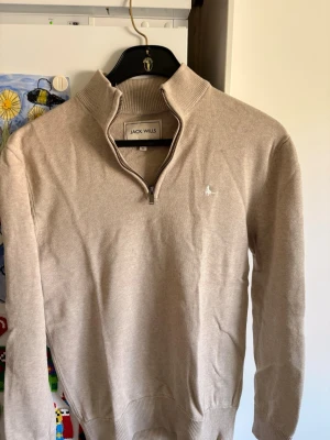 Beige stickad halv zip från Jack Wills - Snygg beige stickad tröja från Jack Wills med halv dragkedja och hög krage. Tröjan har långa ärmar, ribbade muddar och en liten broderad logga på bröstet. Perfekt för lager på lager och enkel att matcha med jeans