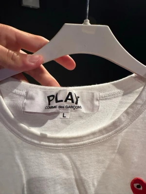 Vit långärmad topp Comme des Garçons - Säljer en vit långärmad topp från Comme des Garçons PLAY i storlek L. Toppen har en klassisk rund halsringning och är tillverkad i mjuk bomull. På bröstet finns det ikoniska röda hjärtat med ögon som ger en unik touch.