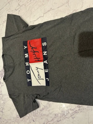 Grå Tommy Jeans t-shirt med tryck - Snygg grå t-shirt från Tommy Jeans med stort tryck framtill i rött, vitt och blått. Klassisk passform med rund hals och korta ärmar. Perfekt för en avslappnad och trendig look.