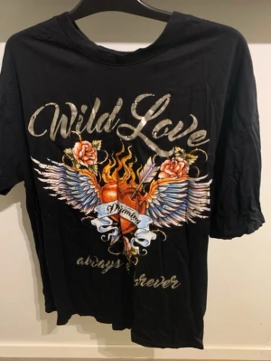 Svart t-shirt med tryck Wild Love - Cool svart t-shirt med stort färgglatt tryck framtill som visar ett brinnande hjärta med vingar, rosor och texten 'Wild Love'. T-shirten har rund hals och korta ärmar. Perfekt för dig som gillar statement-plagg och edgy stil.