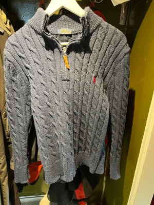 Polo ralph lauren tröja - 8/10 skick. Inget är trasigt eller spruket. Kabelstickad mörkblå tröja från Polo Ralph Lauren med halv dragkedja och hög krage. Tröjan har ett rött broderat logomärke på bröstet och ribbade muddar vid ärmslut och nederkant. Perfekt för dig som gillar klassisk och stilren design. 
