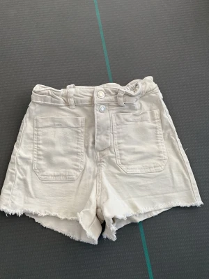 Beiga jeansshorts med rå kant - Säljer ett par beiga jeansshorts med medel hög midja och råa kanter. Shortsen har två stora fickor framtill, bälteshällor och stängs med fyra knappar. Perfekta för sommaren och riktigt snygga till crop tops eller oversized t-shirts.