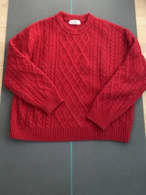 Röd kabelstickad tröja från Lindex - Snygg röd kabelstickad tröja från Lindex med rund halsringning och ribbade muddar. Tröjan har ett klassiskt flätmönster både fram och på ärmarna. Perfekt för dig som vill ha en varm och trendig look i vinter.