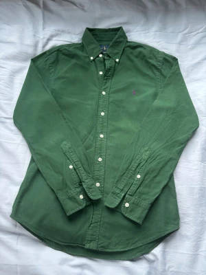 Grön skjorta från Ralph Lauren - Klassisk grön skjorta från Ralph Lauren med lila broderad logga på bröstet. Skjortan har lång ärm, knappar hela vägen och button-down krage. Tillverkad i mjuk bomull som känns skön mot huden. Perfekt för en clean och stilren look.