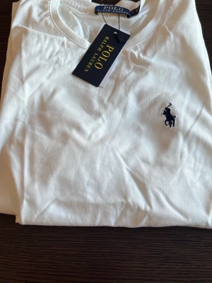 Vit t-shirt från Polo Ralph Lauren - Klassisk vit t-shirt från Polo Ralph Lauren med den ikoniska broderade loggan i marinblått på bröstet. Tillverkad i mjuk bomull och har en normal passform. Perfekt basplagg för en clean och stilren look.