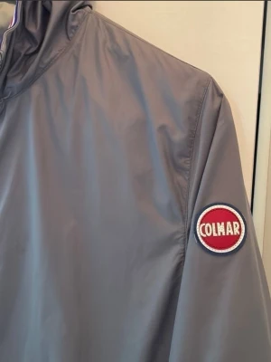 Grå vindjacka med huva från Colmar - Snygg grå vindjacka från Colmar med huva och dragkedja framtill. Jackan har två sidofickor och ett tydligt Colmar-märke på ärmen. Detaljer i rött och vitt vid muddar och krage ger en sportig look. Tillverkad i lätt, vindtätt material som passar perfekt till vardags. Barnjacka, storlek 158,164