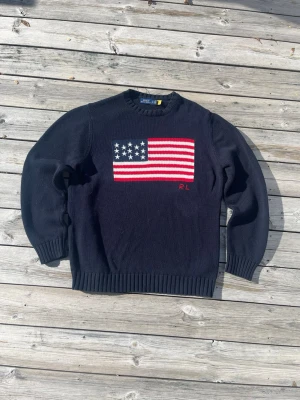 Polo Ralph Lauren stickad tröja - Stickad tröja från Polo Ralph Lauren i mörkblå färg med stor USA-flagga framtill och broderad RL-logo. Klassisk preppy-stil som passar perfekt till både jeans och chinos.  • Märke: Polo Ralph Lauren • Färg: Mörkblå / Navy • Design: Stickad USA-flagga + RL-logo • Skick: Mycket fint skick • Klassisk och tidlös Ralph Lauren-modell