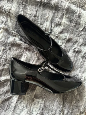 Svarta lackade ballerinaskor med klack - Säljer ett par svarta ballerinaskor i glansigt lackmaterial med T-rem och rund tå. Skorna har en bred, låg klack och spänne vid ankeln för extra detalj. Perfekta för dig som vill ha en klassisk men trendig look.