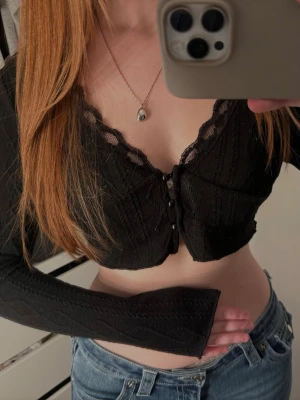 Svart croppad spetsblus - Säljer en svart croppad blus med spetsdetaljer och v-ringad hals. Toppen har långa ärmar och knappar framtill. Perfekt för dig som gillar en feminin och trendig stil.