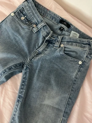 Ljusblå low rise flare jeans från true religion  - Ljusblå jeans med klassisk femficksmodell och utsvängda ben. Jeansen har en något tvättad look och silverfärgade knappar och nitar. Materialet är mjukt denim i bomull, perfekt för en avslappnad stil. Det finns små defekter längst ner som man ser på bilderna men inget annat, använt ungefär tio gånger 