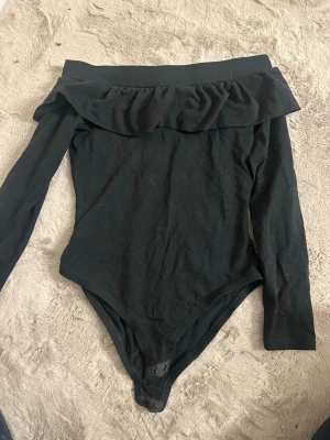 Svart off shoulder body - Säljer en svart body med off shoulder-design och långa ärmar. Bodyn har en bred volang upptill och knäppning i grenen. Perfekt för dig som gillar stilrena och trendiga plagg. Materialet känns mjukt och stretchigt.