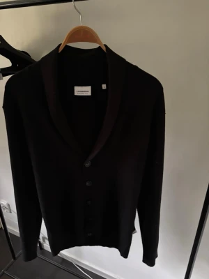 Svart kofta från Lindbergh - Stilren svart kofta från Lindbergh i relaxed fit. Koftan har knäppning framtill med fem knappar och en bred sjalkrage. Perfekt för lager på lager och enkel att matcha med olika outfits.