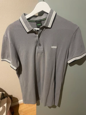 Grå pikétröja från BOSS - Snygg grå pikétröja från BOSS med vit logga på bröstet och vita detaljer på krage och ärmslut. Klassisk polokrage med knappar och korta ärmar. Perfekt till både jeans och shorts för en clean look.