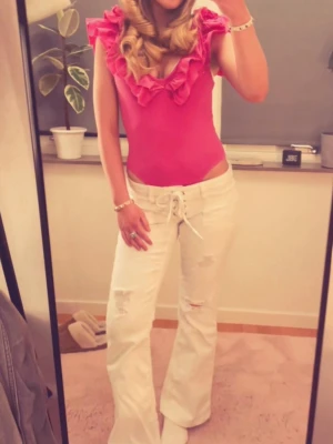 Vita pleated tie jeans - Säljer ett par vita bootcut jeans med snörning i midjan och slitna detaljer på benen. Jeansen har hög midja och vida ben, vilket ger en retro och trendig look. Perfekta för dig som vill sticka ut med en unik stil. 