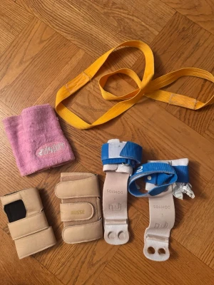 Gymnastiktillbehör Domyos & Russe - Säljer ett kit med gymnastiktillbehör: ett par handskydd i läder från Domyos med blå remmar, handledsskydd från Russe i beige, ett rosa svettband från Allsports och ett gult träningsband. Perfekt för gymnastik eller styrketräning.