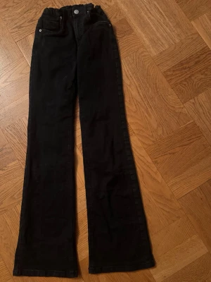 Svarta utsvängda jeans - Säljer ett par svarta jeans med utsvängda ben och hög midja. Klassisk femficksmodell med knapp och dragkedja framtill. Jeansen har en enkel och stilren look, perfekta till en trendig vardagsoutfit. Materialet känns mjukt och följsamt.